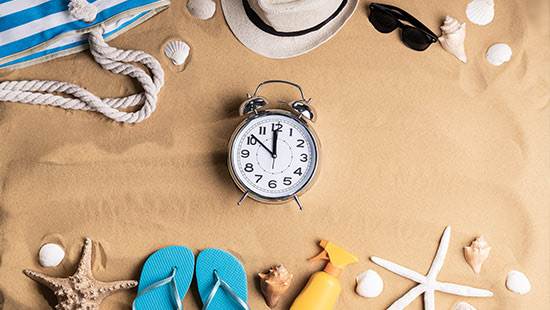 clock on sand surrounded by beach bag, towel, flip flops and sea shells